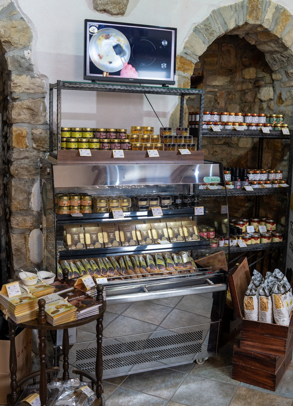 A glass-front refrigerated display case filled with truffle salami, truffle cheeses and butter with truffles. A set in a rustic stone-walled shop with shelves of packaged food gourmet products and a small TV mounted above.